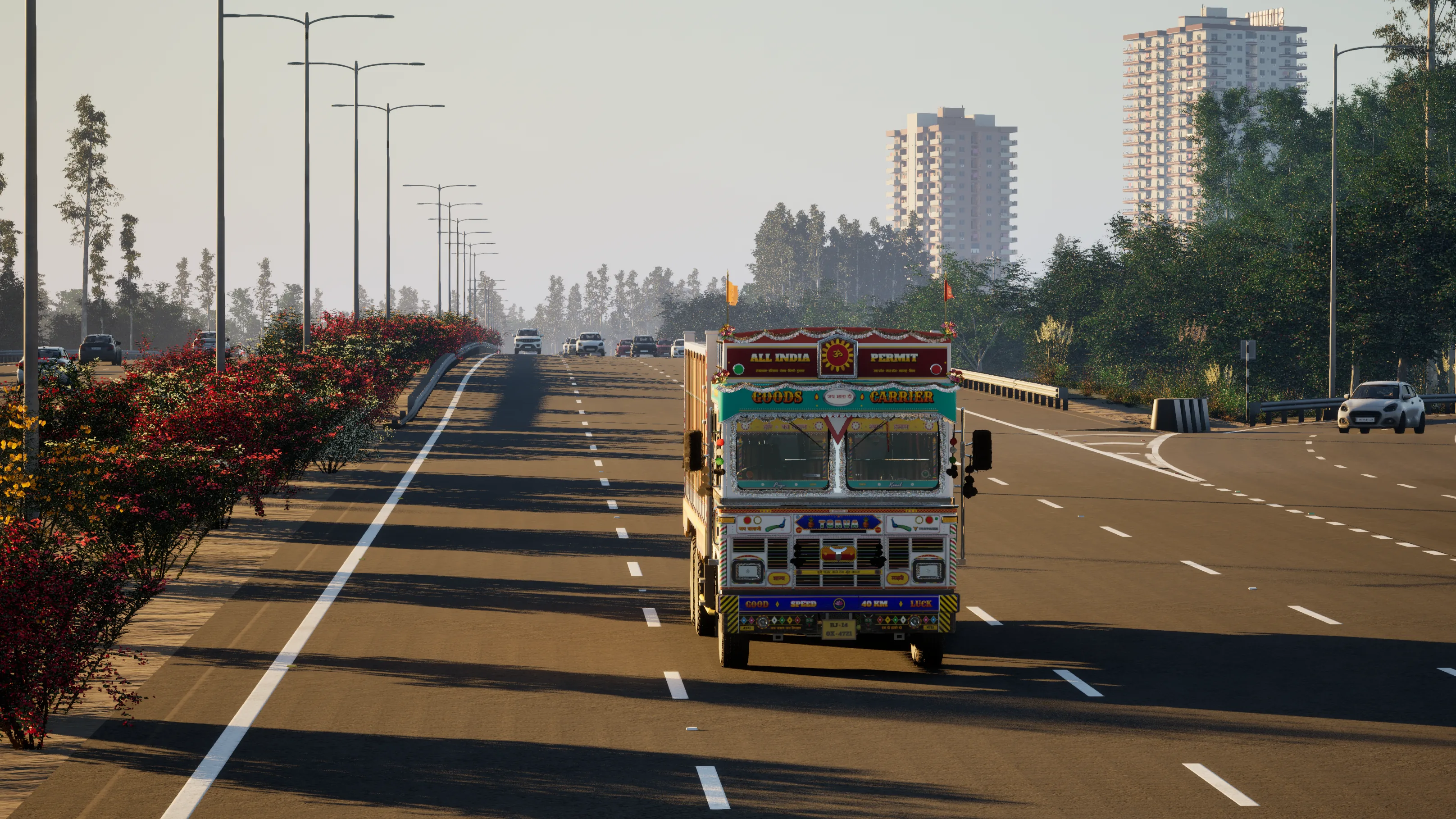 A painted Indian truck driving towards the camera on a wide expressway at golden hour, a flowering median and street lights on the left and apartment towers behind