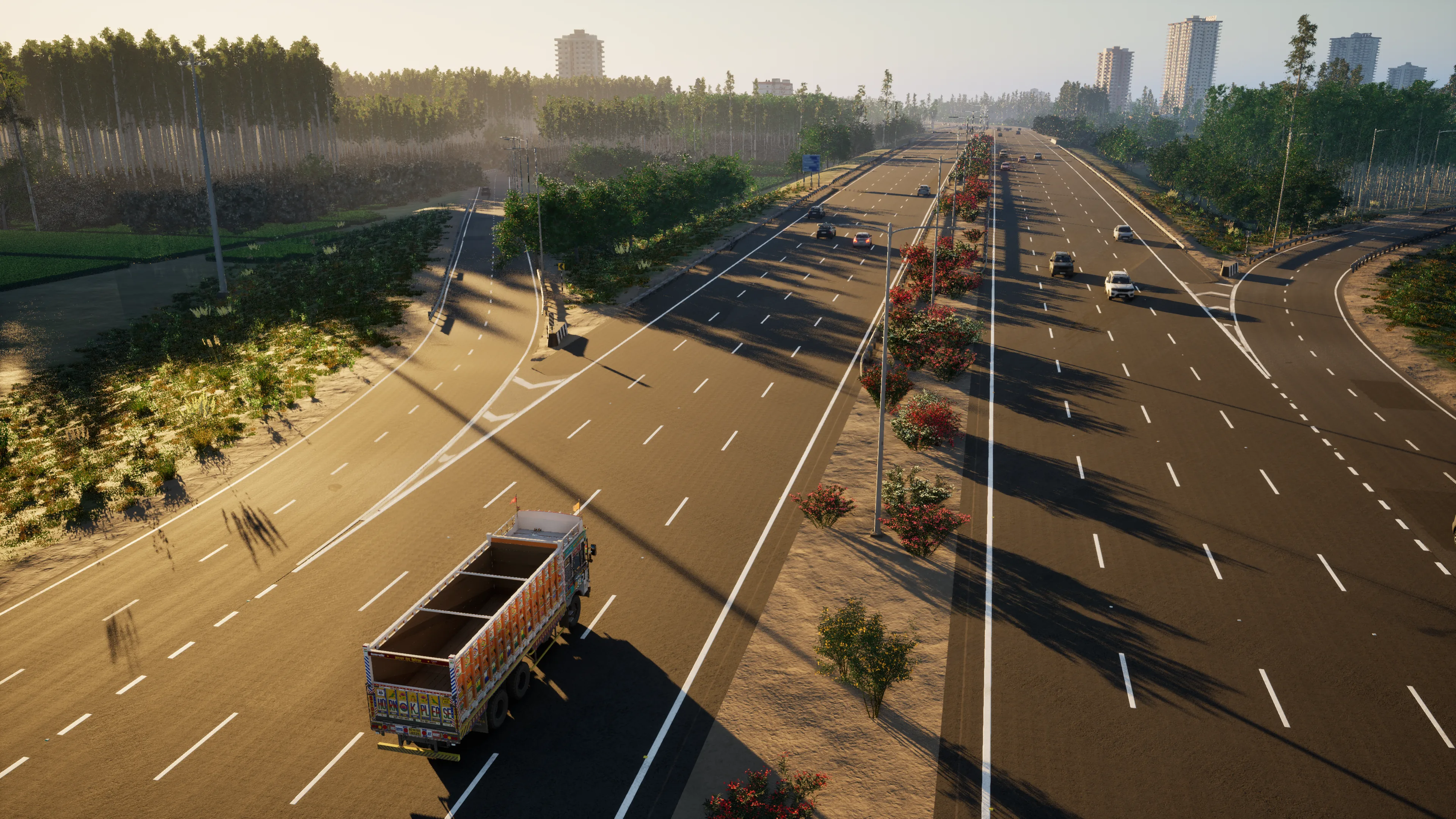 High view of a wide expressway with slip roads and a flowering median at golden hour, a truck in the lower left and towers on the horizon