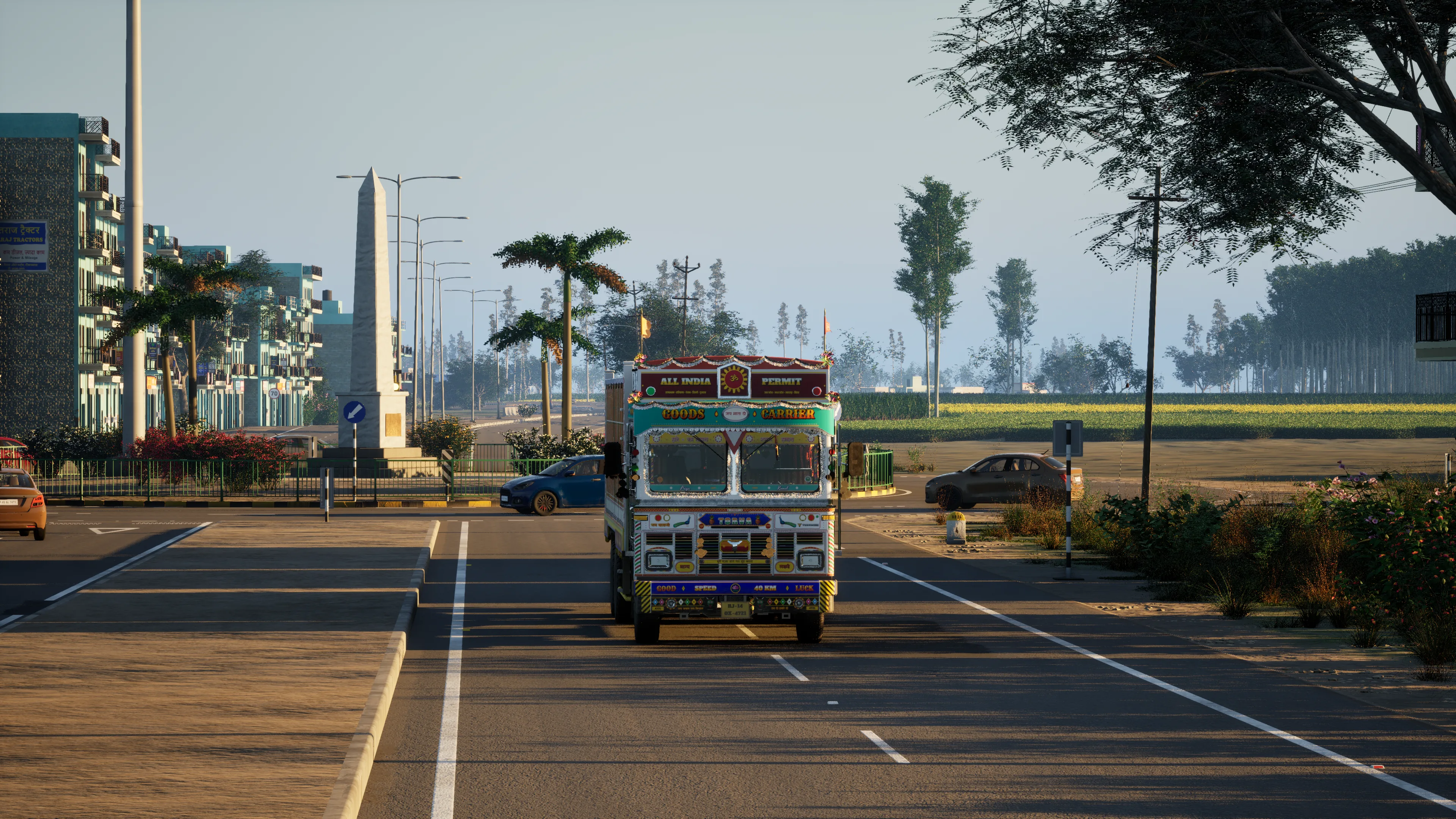 A painted truck leaving a roundabout with a white obelisk and palm trees, flats on the left and a mustard field on the right