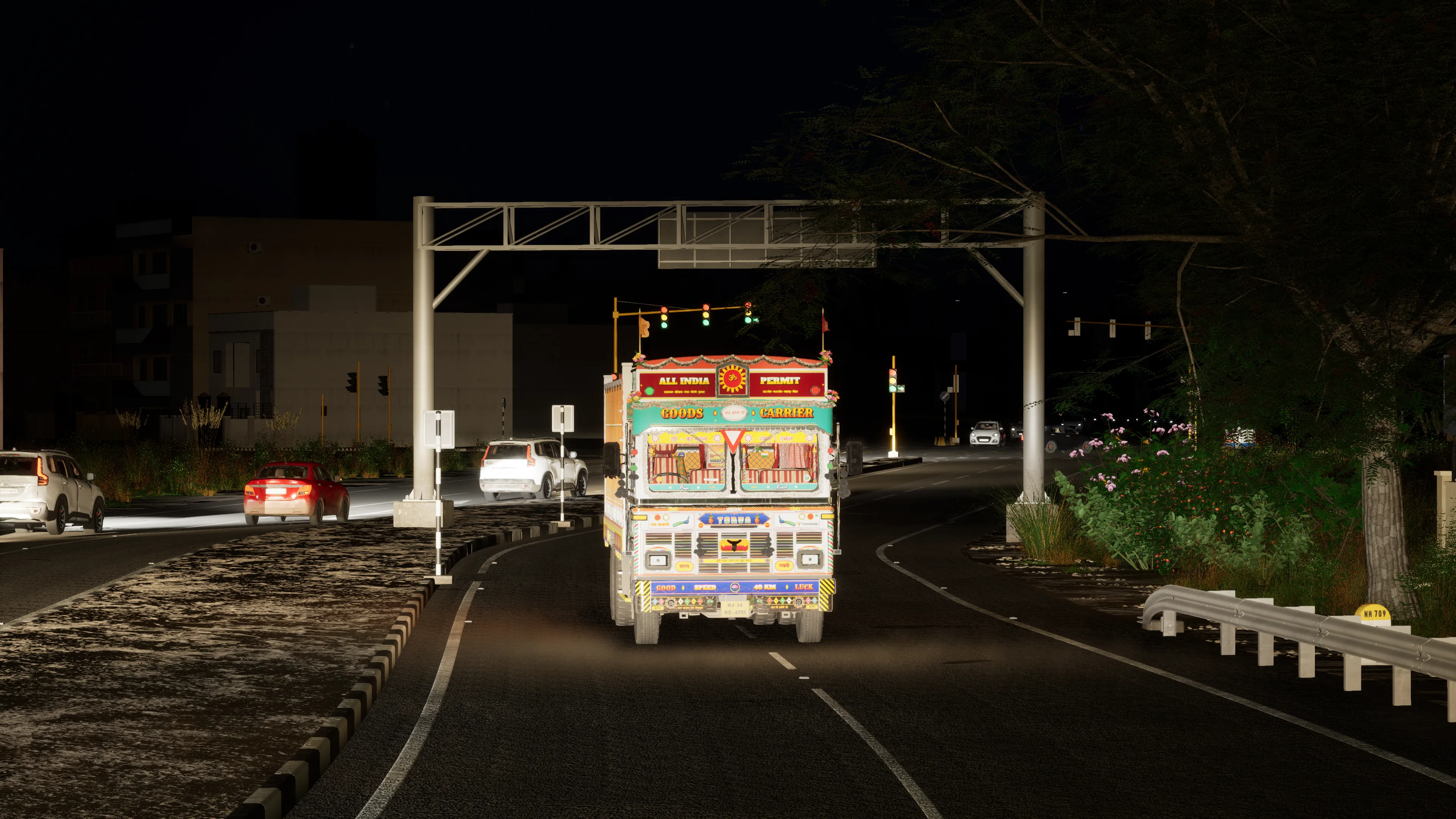 A painted truck lit up at night on a town road, an overhead gantry behind it, traffic lights at the junction beyond and cars on the other carriageway