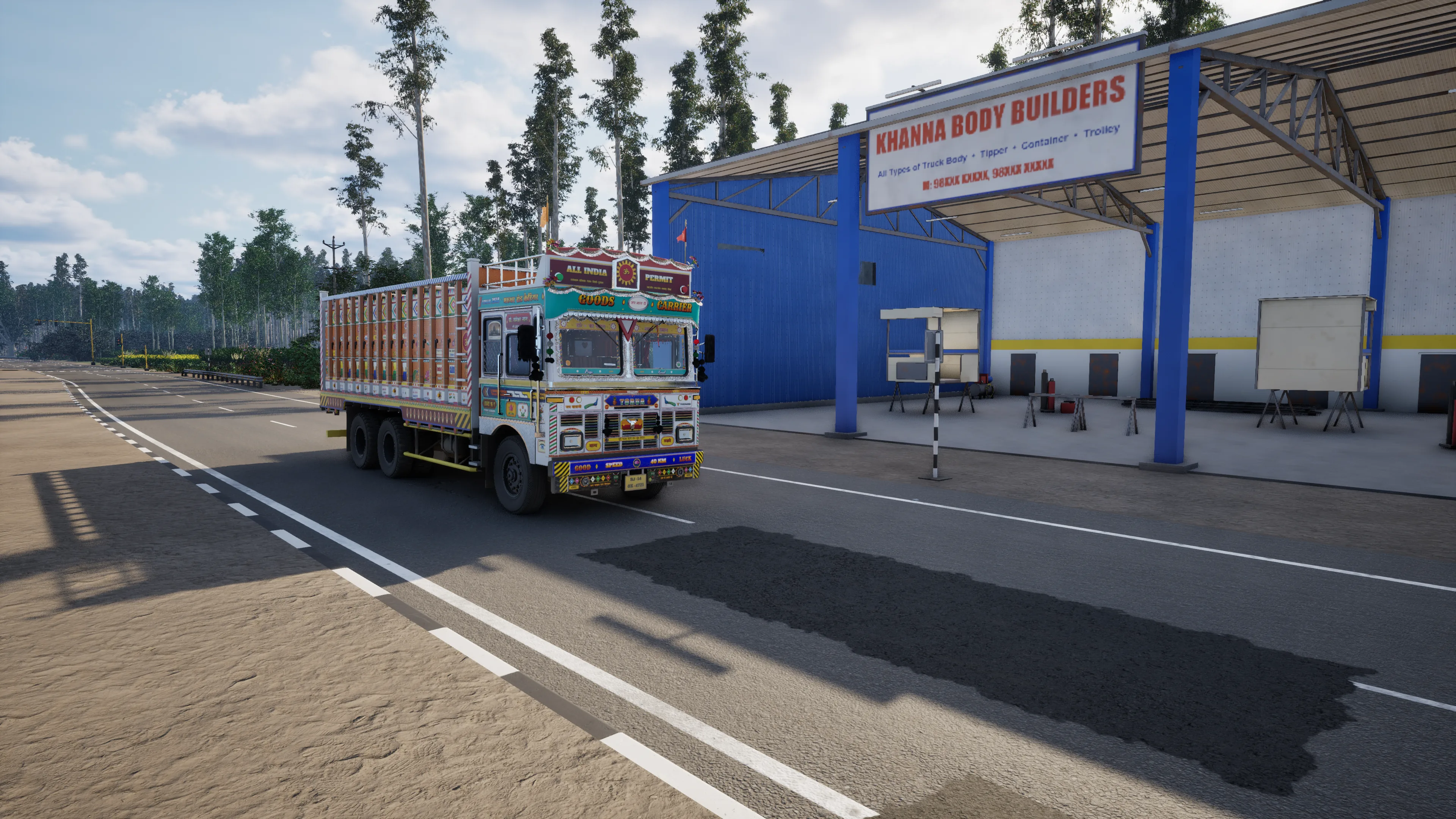 A painted truck in bright sun on a road past the blue shed of a truck-body workshop, a sandy shoulder in the foreground